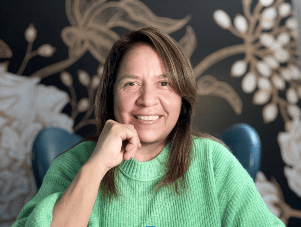 Sonia has dark brown hair that is worn down hitting her shoulders. She is smiling, looking into the camera with brown eyes, wearing a sea green sweater, and hand under chin pose